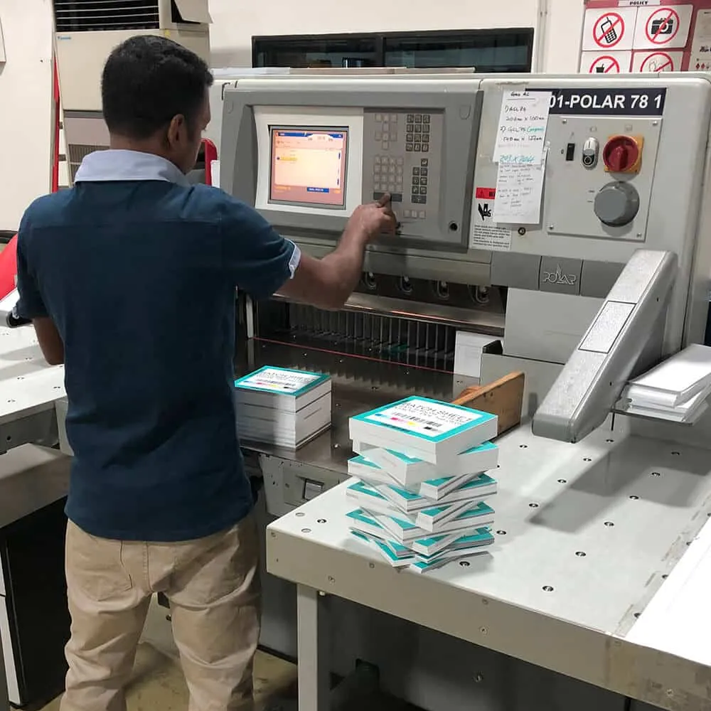 zensmart is showing A man operates a large industrial paper-cutting machine in a workspace. He is wearing a blue shirt and beige pants, pressing buttons on the machine’s screen. Stacks of paper and photo prints are placed next to the machine on a table. The area has various notices posted. with print workflow automation